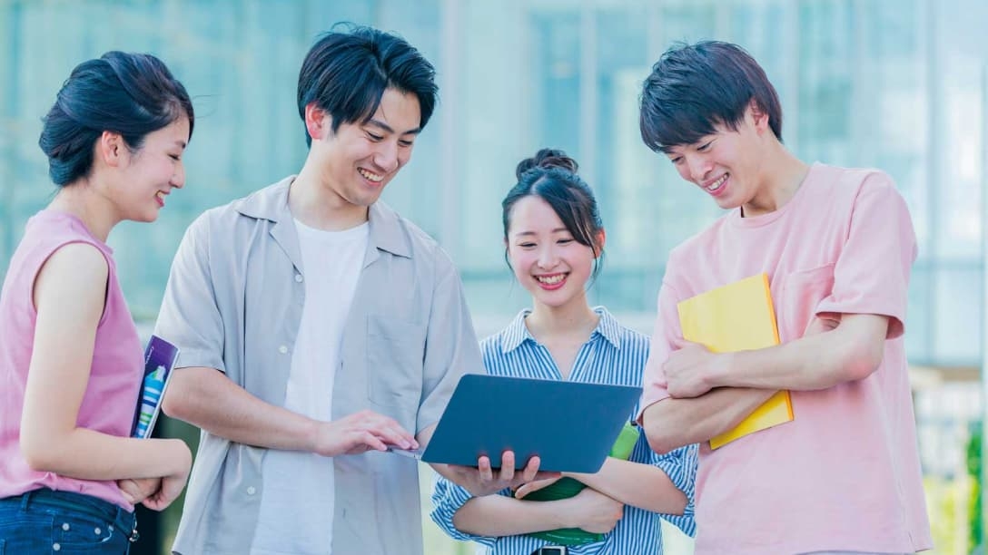 Four smiling students gather around a laptop outdoors